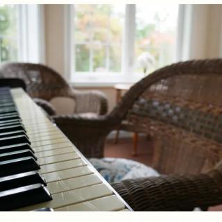 A cozy living room featuring a piano, wicker chairs, and large windows.