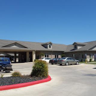 A view of a senior living facility featuring a parking area and well-maintained landscaping under a clear blue sky.