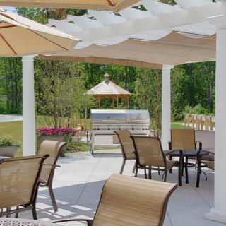 A serene outdoor patio with chairs and tables under a pergola, featuring a grill in the background.