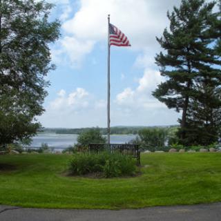 A serene view of an American flag standing tall amidst lush greenery and a scenic backdrop of water and clouds.