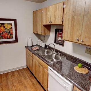A cozy kitchen with wooden cabinets, a granite countertop, a double sink, and a dishwasher, featuring wood flooring and decorative wall art.