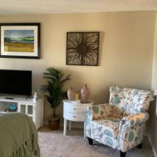 A cozy living room corner featuring a patterned armchair, small side table with vases, TV stand with a television, and decorative artwork on the walls.