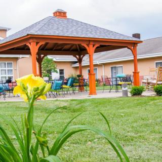 A sunny garden with a gazebo and outdoor seating surrounded by a lush lawn and blooming flowers.
