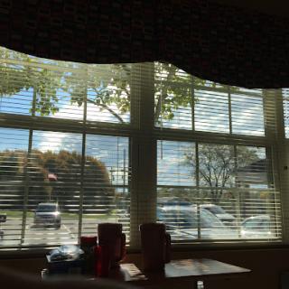A view through a window with blinds, showcasing trees and a partly cloudy sky.