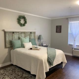 A cozy bedroom with soft green and white decor, featuring a bed with pillows, wall art, and a window letting in natural light.