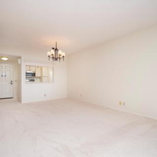 An empty living room with white walls and carpet, featuring a chandelier and a small kitchenette area.