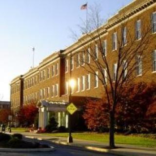 A large brick building with numerous windows, surrounded by autumn trees along a road.
