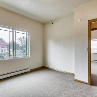 An empty room in an apartment with a large window, carpeted floors, and white walls.