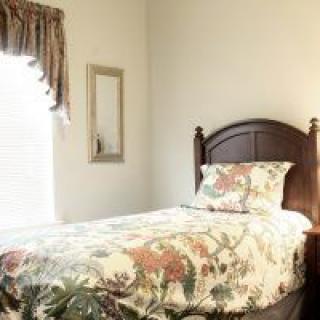 A cozy bedroom with a single bed featuring a floral bedspread, accompanied by a window with curtains and a wall mirror.