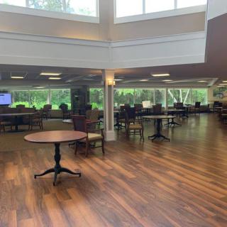 An empty room with wooden flooring, tables, and chairs, illuminated by natural light from large windows.