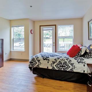 A cozy bedroom with wooden flooring, featuring a bed adorned with patterned bedding and bright throw pillows, illuminated by natural light from large windows.