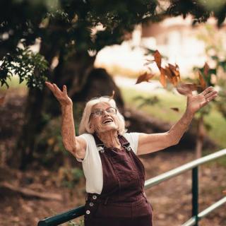An elderly woman joyfully throws autumn leaves into the air while enjoying the outdoors.
