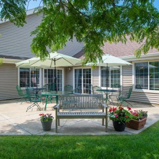 A cozy outdoor patio with garden furniture, including a bench and tables under umbrellas, surrounded by greenery.