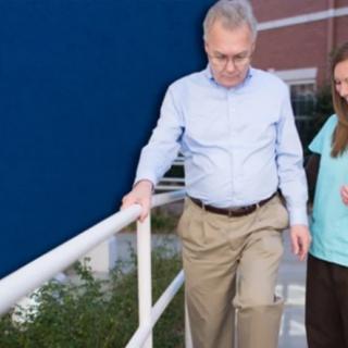 An elderly man receives assistance from a nurse while walking outdoors.
