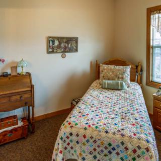 A cozy bedroom with a single bed featuring a colorful quilt, wooden desk, and window.