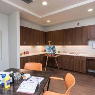 A cozy art room featuring an easel with a painting, surrounded by wooden cabinets and a table with chairs.