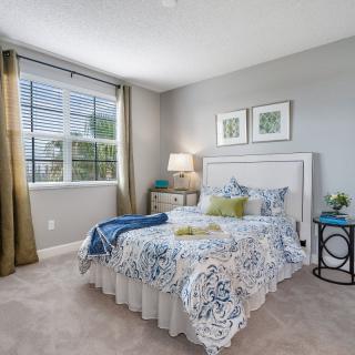 A well-lit bedroom with a patterned bedspread, a bedside lamp, and framed artwork on the wall.