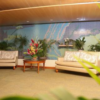 A cozy lobby area with two sofas and a coffee table adorned with flowers, surrounded by indoor plants and a mural artwork on the wall.