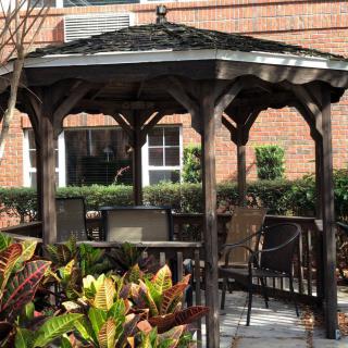 A wooden gazebo with chairs and tables, surrounded by plants outside a brick building.