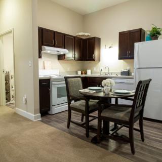 A modern apartment kitchen with a dining area featuring a table and chairs, dark wooden cabinets, and white appliances.