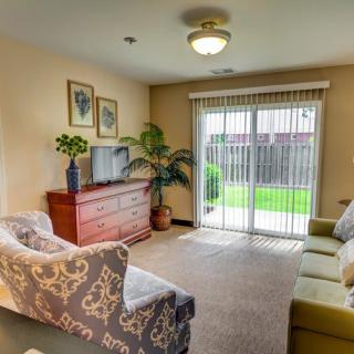 A cozy living room with a sofa, armchair, dresser, television, and indoor plants, featuring a sliding glass door leading to an outdoor area.