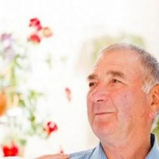 An older man smiling indoors with flowers in the background.