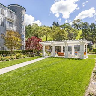 A scenic view of a manicured lawn with a pergola, adjacent to a building, set against a backdrop of lush trees and a bright, cloudy sky.