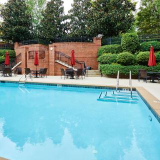An inviting outdoor swimming pool surrounded by chairs, tables, and red umbrellas, with lush greenery in the background.