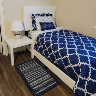A cozy bedroom with a single bed featuring blue patterned bedding, a white nightstand, and a decorative lamp on a wooden floor.