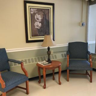 A cozy waiting area featuring two blue chairs, a small wooden table with a lamp, and framed artwork on the wall.