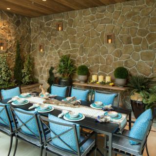 An elegant outdoor dining area with a stone wall backdrop, featuring blue cushioned chairs and a well-set table surrounded by lush greenery.