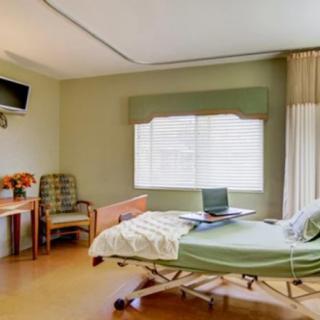 A well-lit hospital room featuring a bed, a chair, and a small desk with flowers.