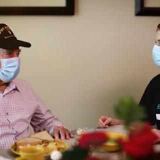 An elderly man and a caregiver having a conversation over a meal while wearing masks.
