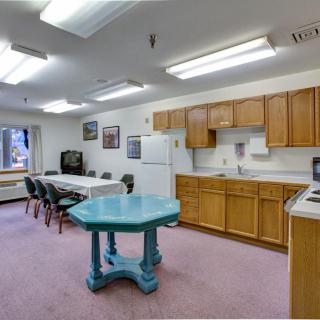 A community room with a kitchen setup, featuring wood cabinets, a refrigerator, tables, and chairs.