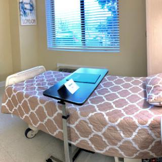A patient room featuring a hospital bed with a blanket, a bedside table, and natural light coming through the window.