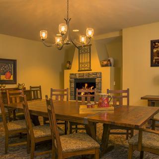 A cozy dining room with a wooden table and chairs, illuminated by a chandelier above and featuring a fireplace in the background.