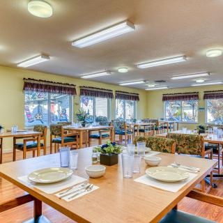 A spacious dining room with neatly arranged tables and chairs, illuminated by ceiling lights and natural daylight from large windows.