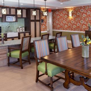 A cozy dining area with a large wooden table, cushioned chairs, and warm lighting, featuring floral wallpaper in the background.
