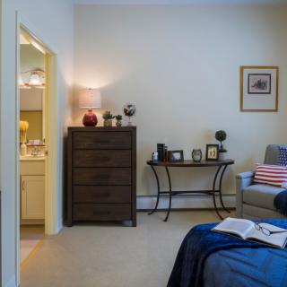 A cozy bedroom with a dresser, table, and comfortable chair, decorated with lamps and framed pictures.