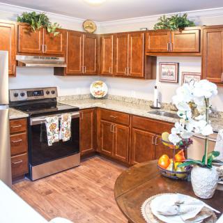 A modern kitchen with wooden cabinets and stainless steel appliances, featuring a corner layout and a small dining area adorned with plants and decorative items.