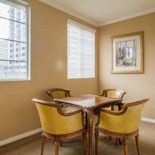 A cozy dining area with a wooden table and four chairs, featuring large windows and a framed painting on the wall.