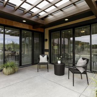 A modern outdoor patio with sleek chairs and a glass door entrance, featuring a pergola overhead and decorative plants.