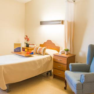 A cozy and well-lit hospital or care facility room featuring a bed, an armchair, and a nightstand.