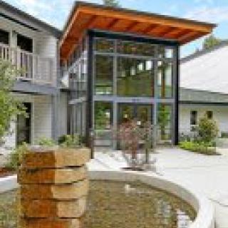 A modern architectural building featuring large glass windows, a wooden roof overhang, and a stone water feature in a courtyard.