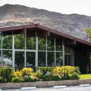 A modern building with large glass windows set against a mountainous backdrop, surrounded by lush greenery.