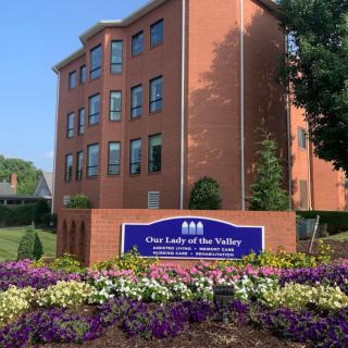 A brick building with a sign reading 'Our Lady of the Valley' surrounded by colorful flowers.