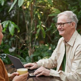 Two elderly individuals enjoying a conversation over coffee while using a laptop in a lush outdoor setting.