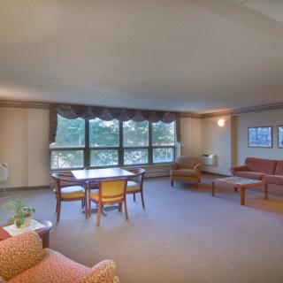 A spacious living room with large windows, several chairs and tables on a carpeted floor.