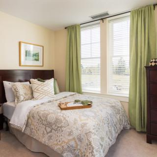 A cozy bedroom with a neatly made bed and green curtains letting in natural light through the windows.