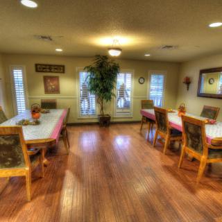 A cozy dining room with wooden floors, decorated with tables and chairs, plants, and ample natural light from large windows.